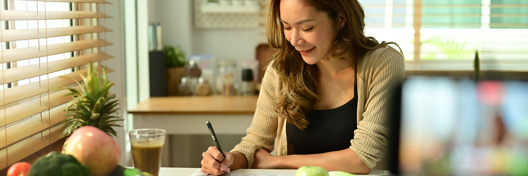 Woman writing at kitchen table surrounded by fruits and vegetables 