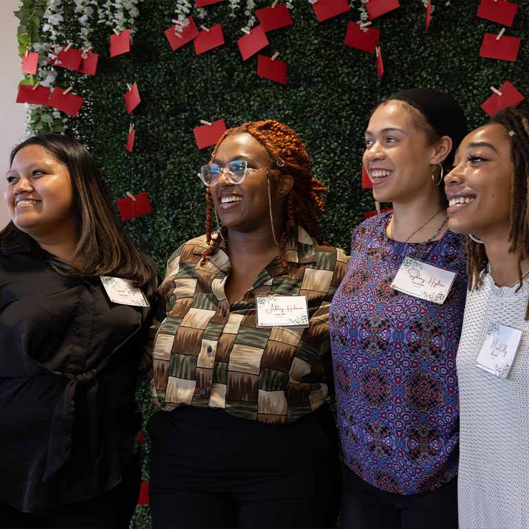 Four women pose in front of a wall decorated with green leaves and red cards while at a scholarship event