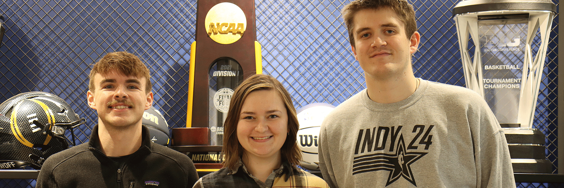 Three sport management students from IU Indianapolis pose for a photo in front of the NCAA Division 1 National Champions trophy. 